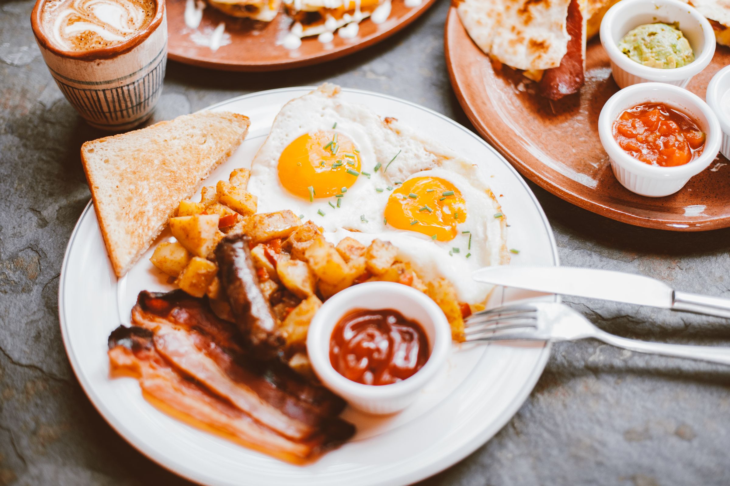 Photograph of a diner breakfast: fried eggs, hash browns, bacon and toast with a coffee alongside.